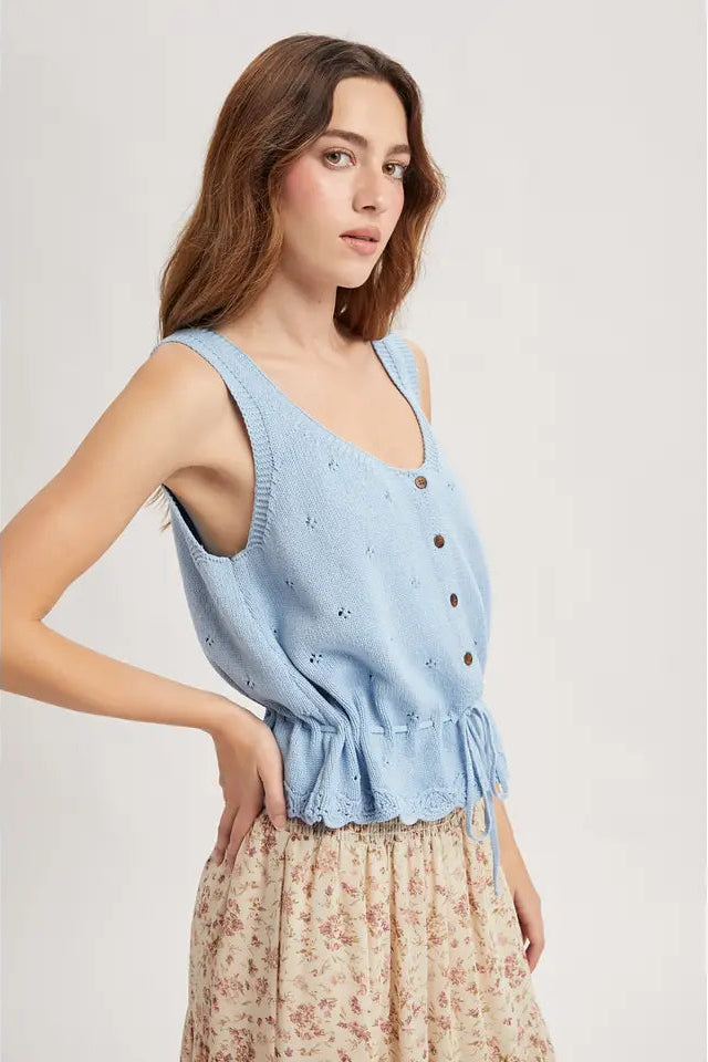 Woman wearing a sky blue sleeveless knitted tank top with buttons and scallop hem on a plain background
