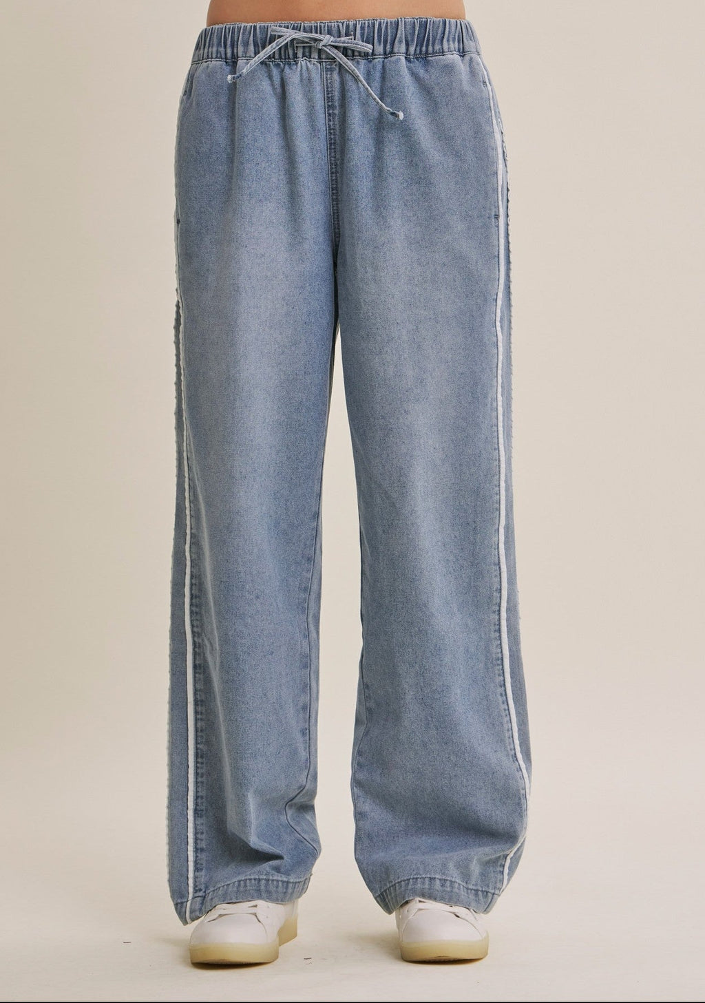 A woman wearing light blue wide-leg denim track pants with a drawstring waist and white sneakers, shown from the waist down against a plain beige studio background.