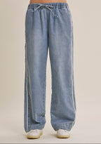 A woman wearing light blue wide-leg denim track pants with a drawstring waist and white sneakers, shown from the waist down against a plain beige studio background.