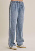 A woman wearing light blue wide-leg denim track pants with a drawstring waist and white sneakers, shown from the waist down against a plain beige studio background.