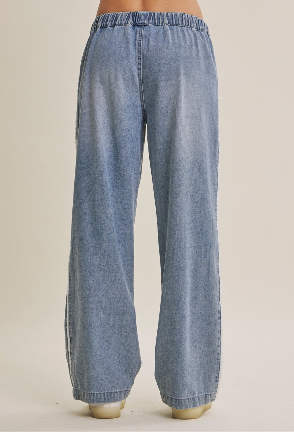 Wide-leg light blue cotton denim track pants with an elastic waistband, shown from the back on a model against a warm beige studio background.