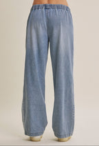 Wide-leg light blue cotton denim track pants with an elastic waistband, shown from the back on a model against a warm beige studio background.