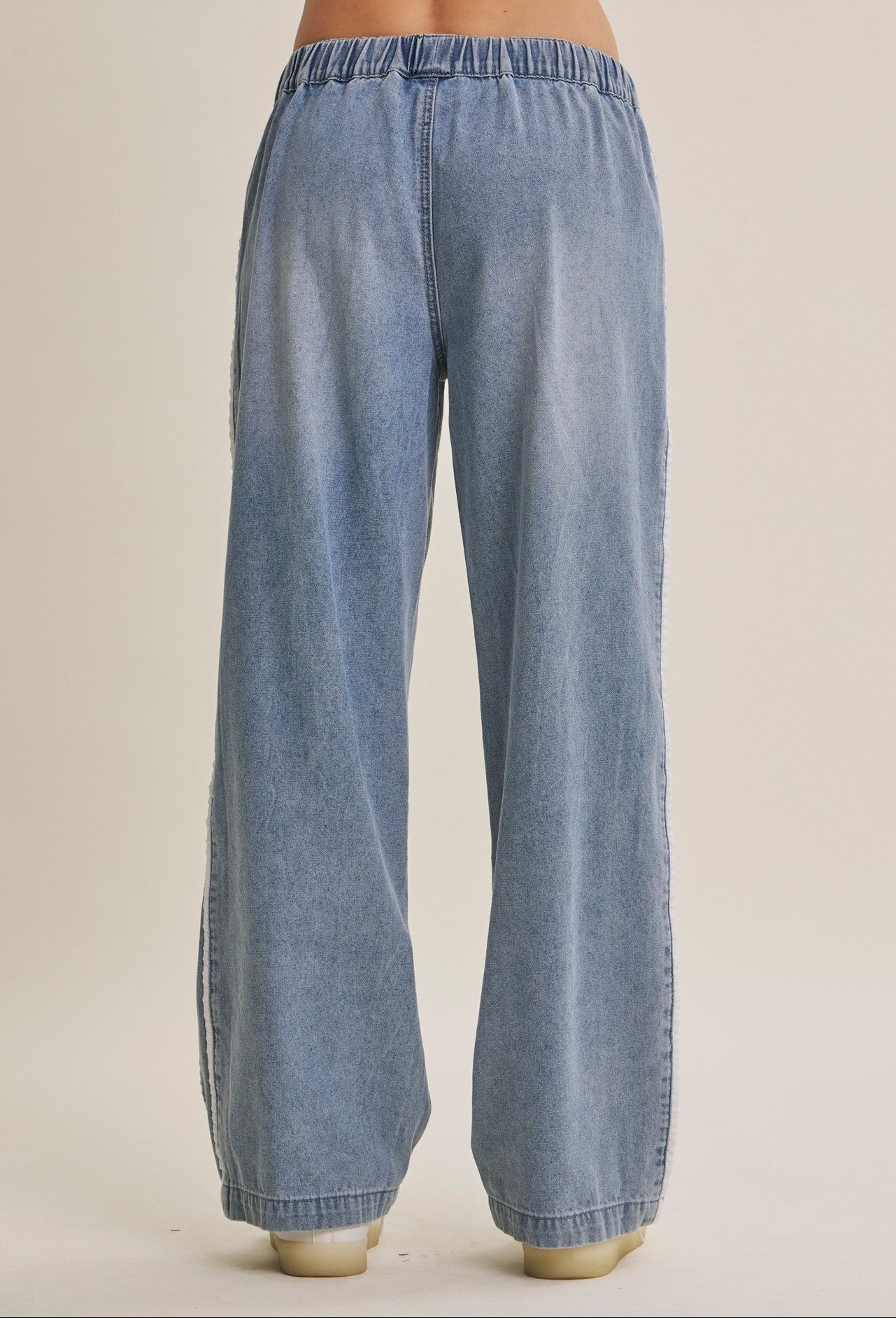 Wide-leg light blue cotton denim track pants with an elastic waistband, shown from the back on a model against a warm beige studio background.
