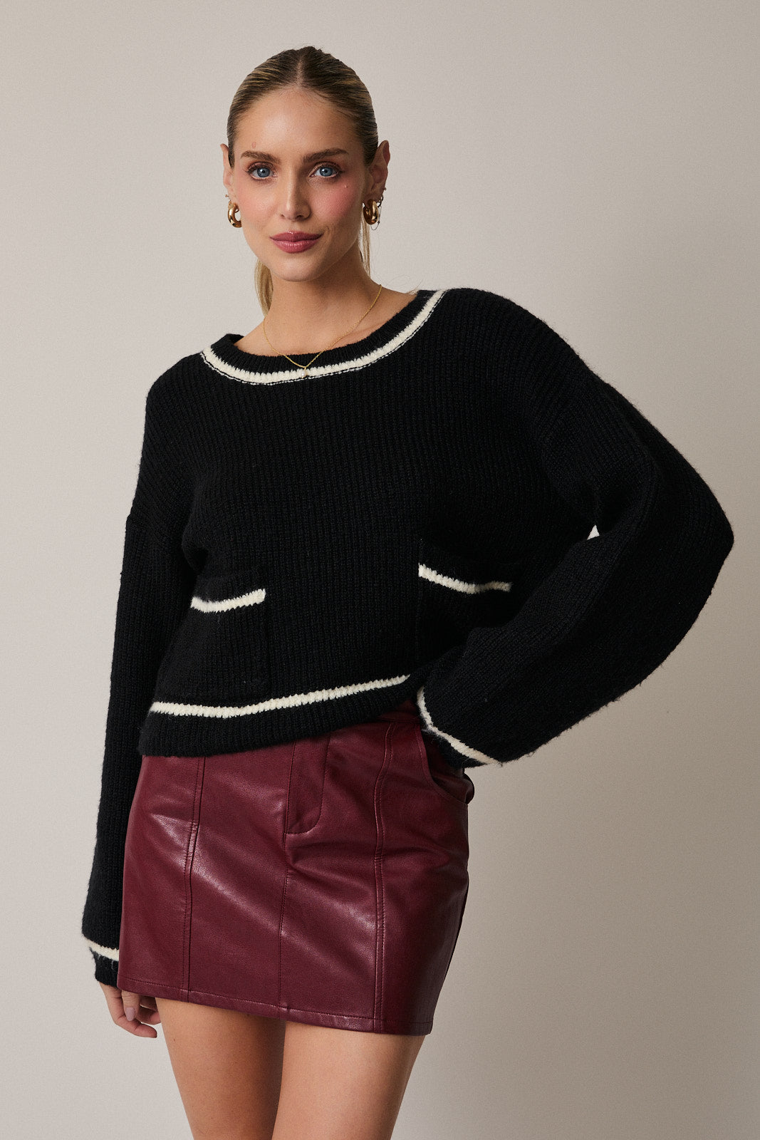 Woman wearing a black sweater with cream trim and a maroon leather skirt on a plain background