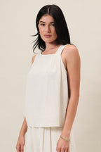Woman wearing a natural linen blend sleeveless top with one side pleat detail against a white background