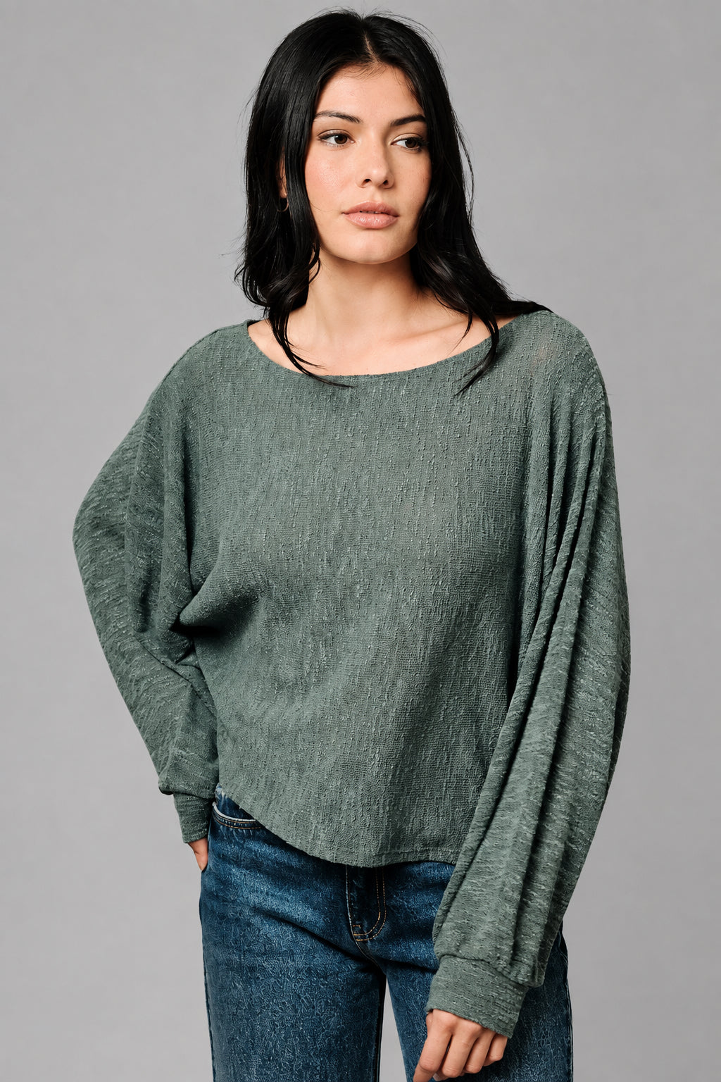 A woman wearing a loose, long-sleeve hunter green top and blue jeans, standing against a textured beige studio backdrop.
