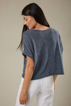 Back view of a woman wearing an indigo knit top and white pants against a beige background