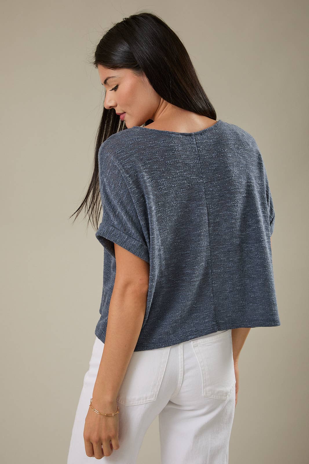 Back view of a woman wearing an indigo knit top and white pants against a beige background