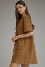 A woman modeling a brown suede-like short-sleeve shift dress with large front patch pockets and decorative metal hardware, photographed against a plain light gray studio background.