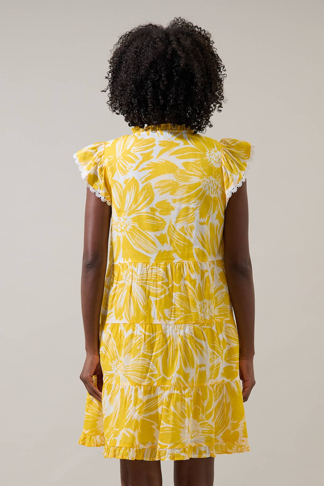 Back vieww of a woman wearing a yellow floral dress with flutter sleeves against a plain background