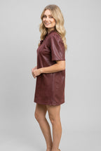 A woman modeling a short-sleeve, v-neck with ruffle dress in a burgundy faux leather fabric, set against a gray background