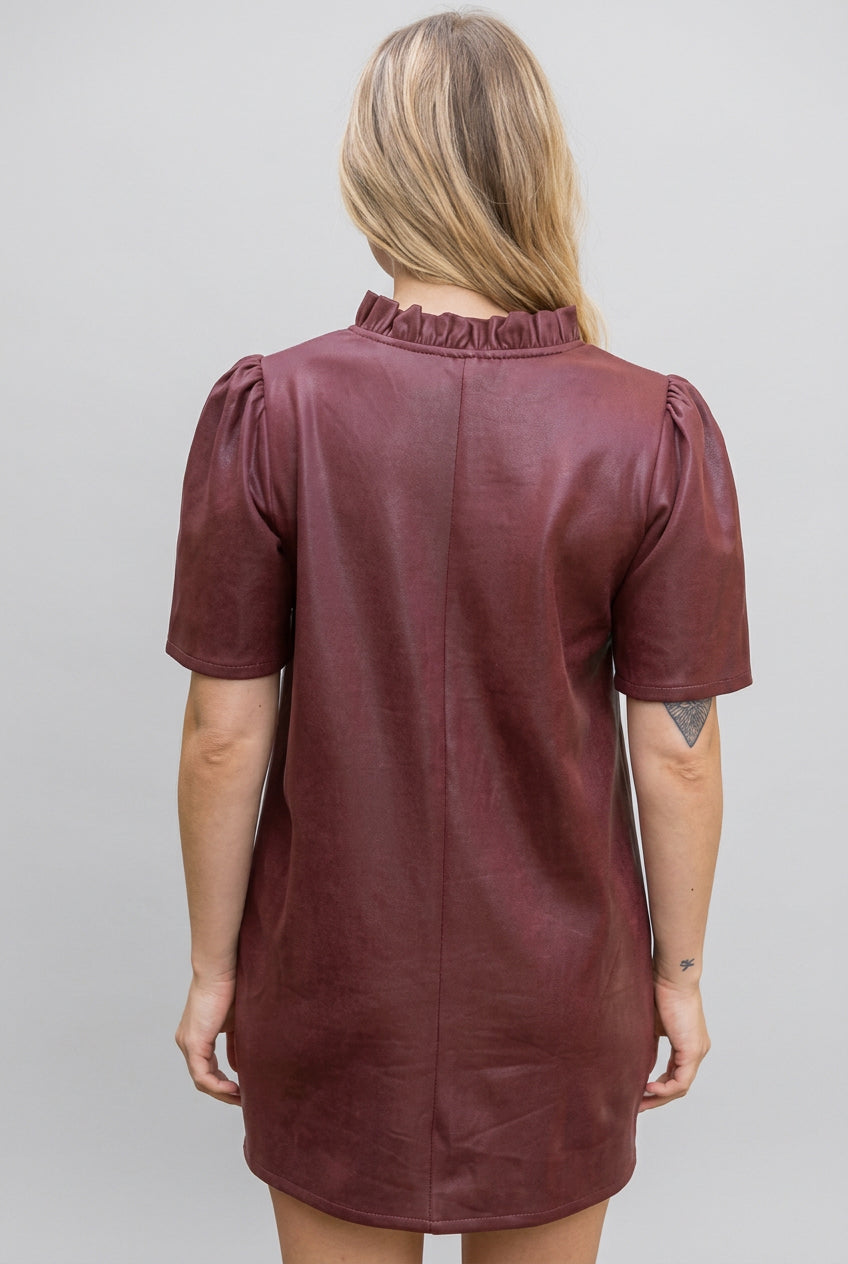Back view of a woman modeling a short-sleeve, v-neck with ruffle dress in a burgundy faux leather fabric, set against a gray background