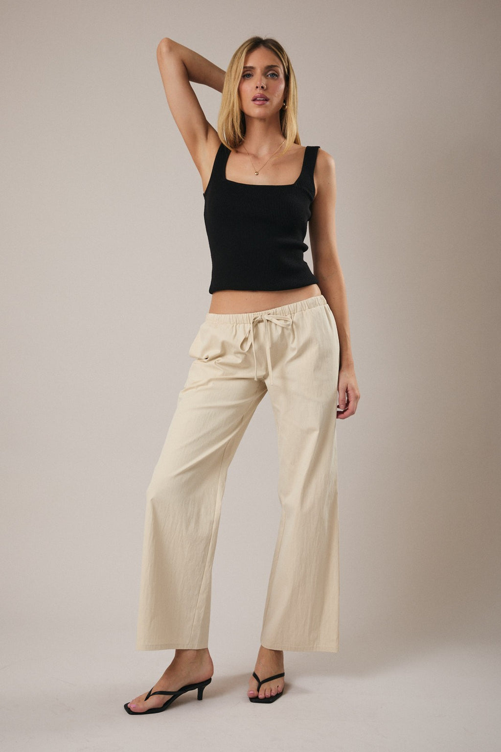 Woman wearing straight leg ecru drawstring pants and black tank top against a grey background