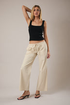 Woman wearing straight leg ecru drawstring pants and black tank top against a grey background