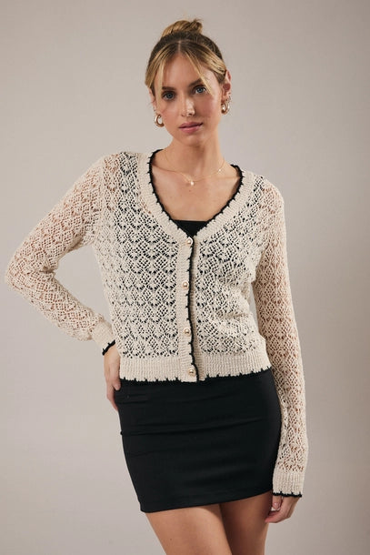 Woman wearing a cream crochet cardigan with black trim over a black top and skirt on a neutral background