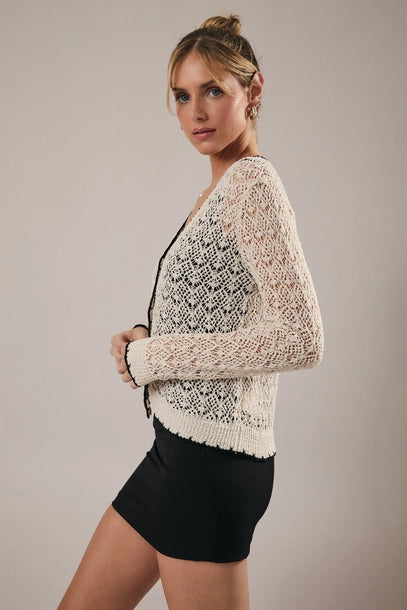 Woman wearing a cream crochet cardigan with black trim over a black top and skirt on a neutral background