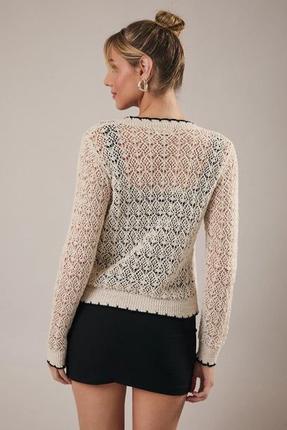 Back view of a woman wearing a cream crochet cardigan with black trim over a black top and skirt on a neutral background