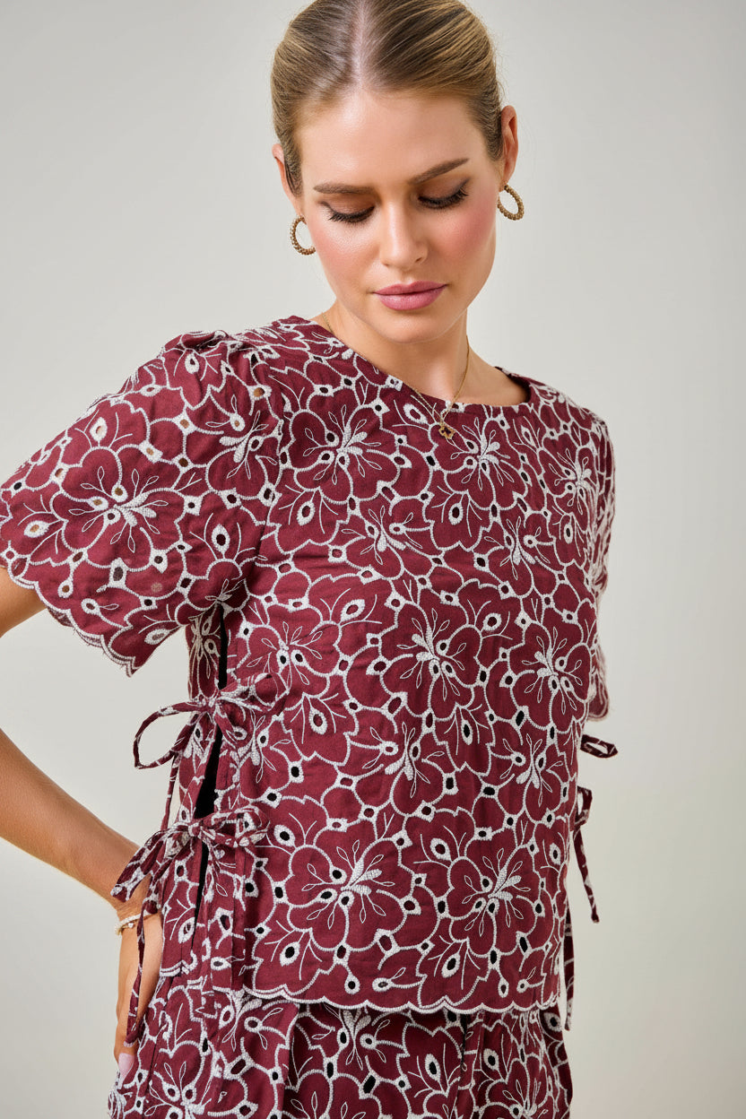 A woman wearing a maroon and white floral embroidered eyelet top with short sleeves and side tie details against a plain beige background.