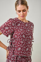 A woman wearing a maroon and white floral embroidered eyelet top with short sleeves and side tie details against a plain beige background.