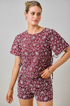 A woman wearing a maroon and white floral embroidered eyelet top with short sleeves and side tie details with matching shorts against a plain beige background. 