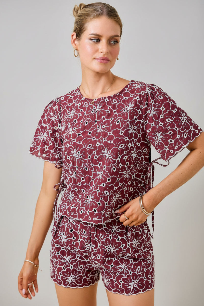 A woman wearing a maroon and white floral embroidered eyelet top with short sleeves and side tie details with matching shorts against a plain beige background. 