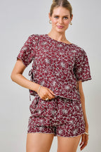 A woman wearing a maroon and white floral embroidered eyelet top with short sleeves and side tie details with matching shorts against a plain beige background.