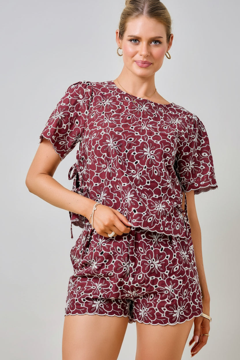 A woman wearing a maroon and white floral embroidered eyelet top with short sleeves and side tie details with matching shorts against a plain beige background.