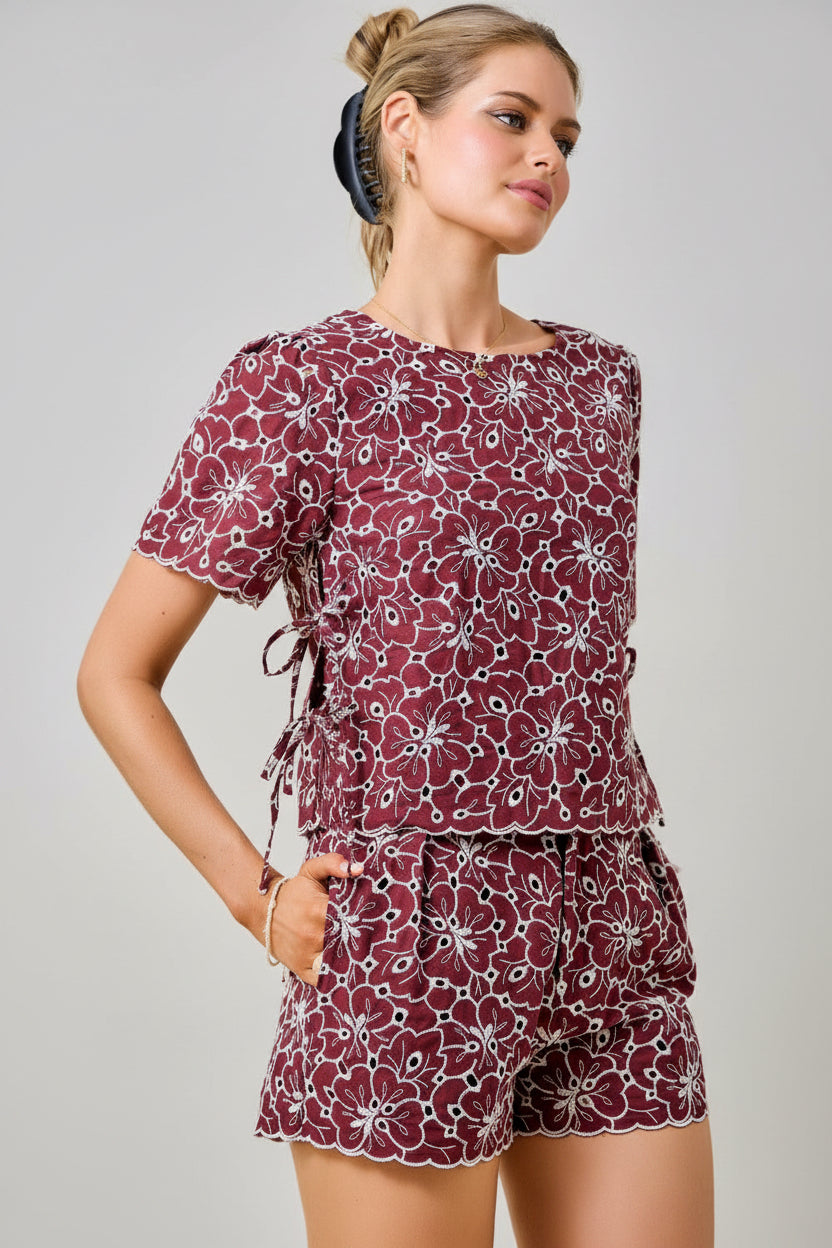 A woman wearing a maroon and white floral embroidered eyelet top with short sleeves and side tie details with matching shorts against a plain beige background.