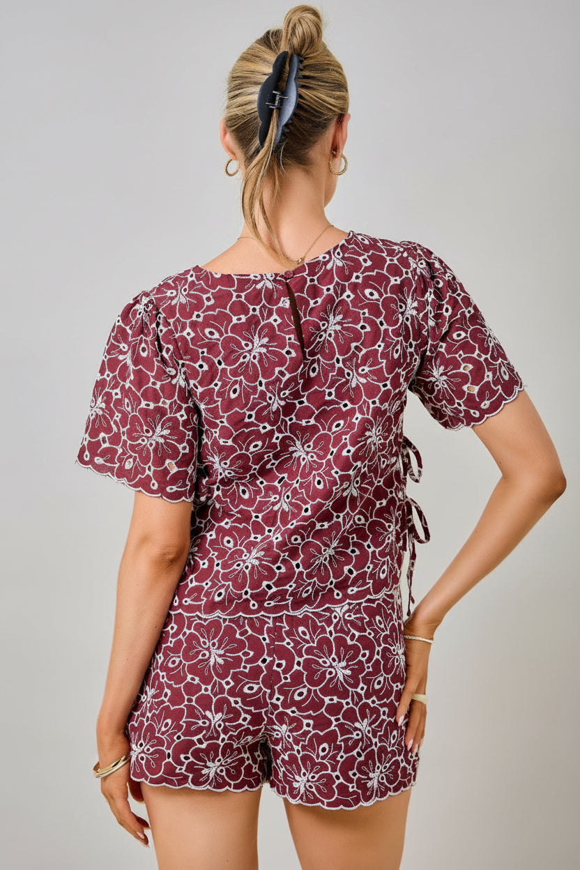 Back view of a woman wearing a maroon and white floral embroidered eyelet top with short sleeves and side tie details with matching shorts against a plain beige background.