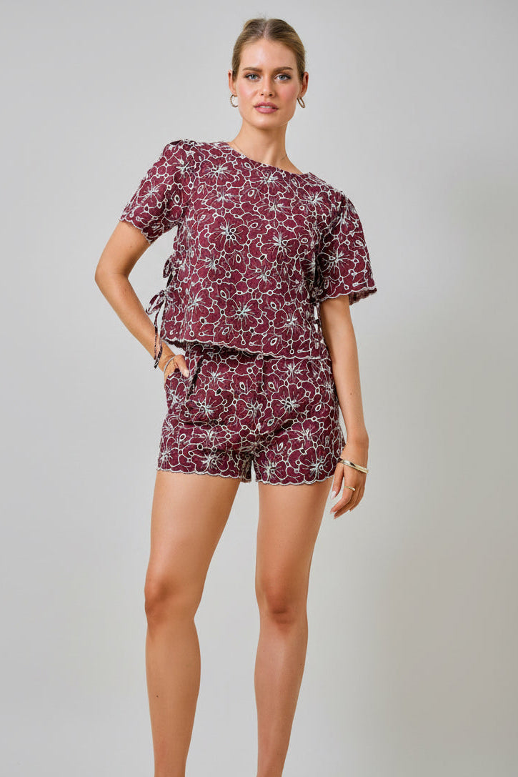 A woman wearing a maroon and white floral embroidered eyelet top with short sleeves and side tie details with matching shorts against a plain beige background.