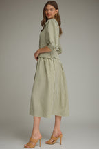A woman modeling an olive green and white vertically striped midi dress with three-quarter sleeves and a tie waist and drop waist, paired with tan heeled sandals against a neutral gray studio background.