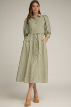 A woman modeling an olive green and white vertically striped midi dress with three-quarter sleeves and a tie waist and drop waist, paired with tan heeled sandals against a neutral gray studio background.