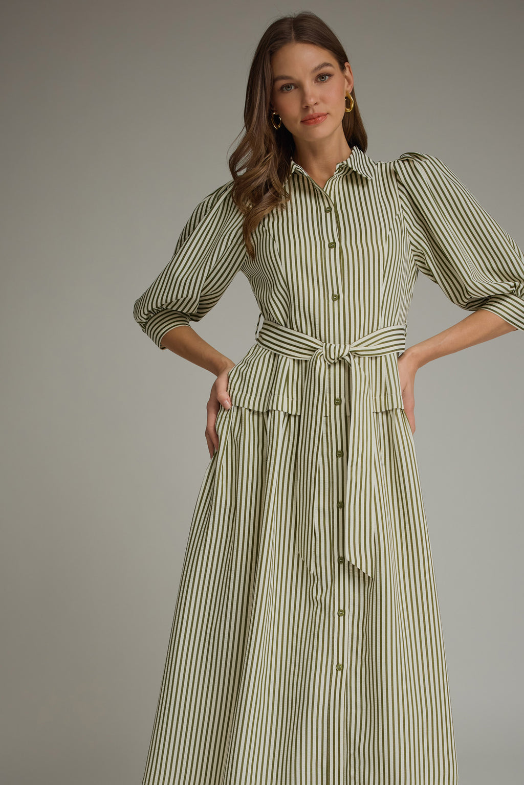 A woman modeling an olive green and white vertically striped midi dress with three-quarter sleeves and a tie waist and drop waist, paired with tan heeled sandals against a neutral gray studio background.