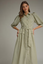 A woman modeling an olive green and white vertically striped midi dress with three-quarter sleeves and a tie waist and drop waist, paired with tan heeled sandals against a neutral gray studio background.
