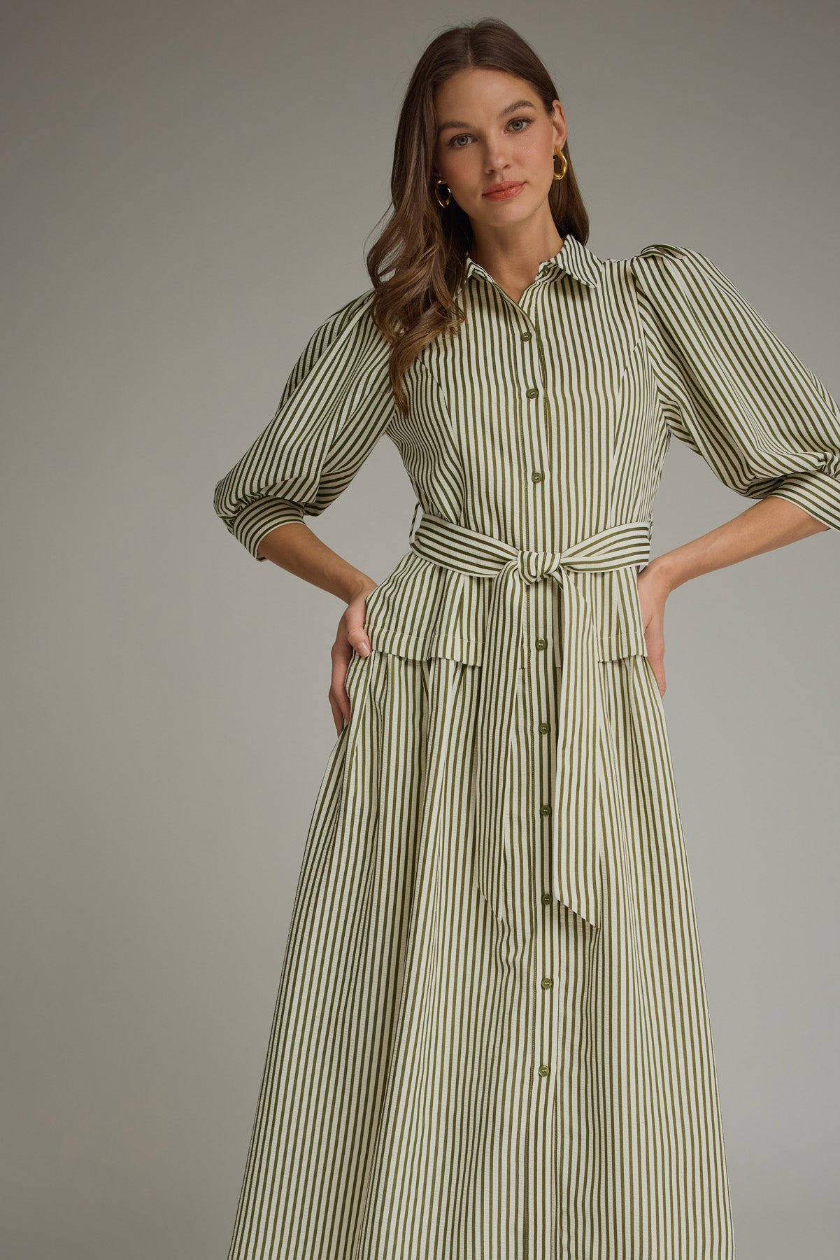 A woman modeling an olive green and white vertically striped midi dress with three-quarter sleeves and a tie waist and drop waist, paired with tan heeled sandals against a neutral gray studio background.