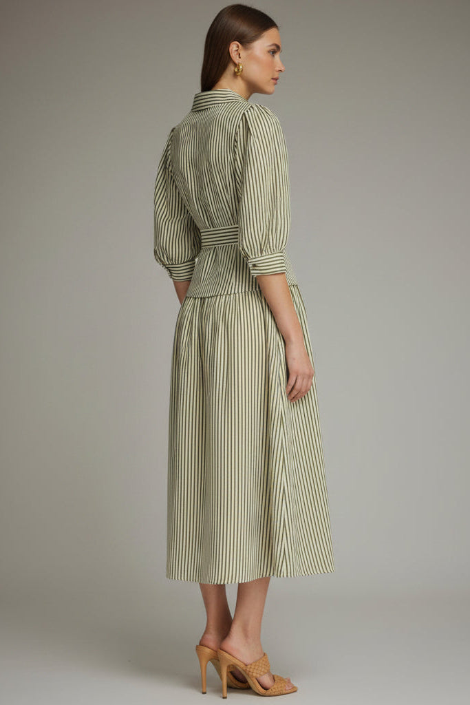 Back view of a woman modeling an olive green and white vertically striped midi dress with three-quarter sleeves and a tie waist and drop waist, paired with tan heeled sandals against a neutral gray studio background.
