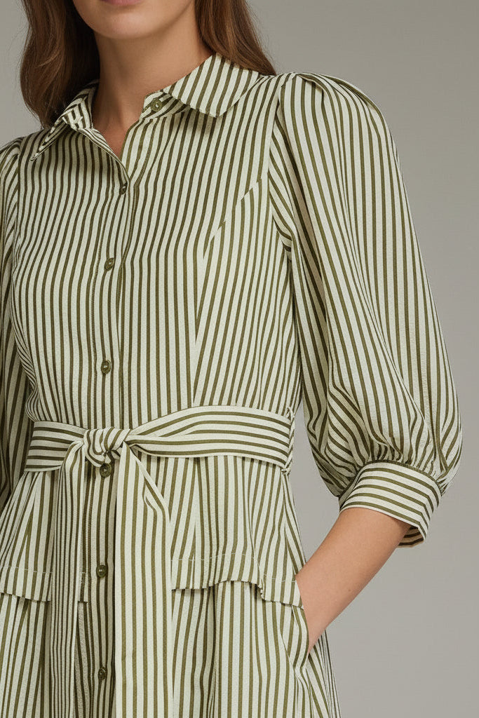 A woman modeling an olive green and white vertically striped midi dress with three-quarter sleeves and a tie waist and drop waist, against a neutral gray studio background.