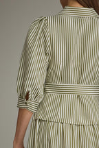 Back view of a woman modeling an olive green and white vertically striped midi dress with three-quarter sleeves and a tie waist and drop waist, against a neutral gray studio background.