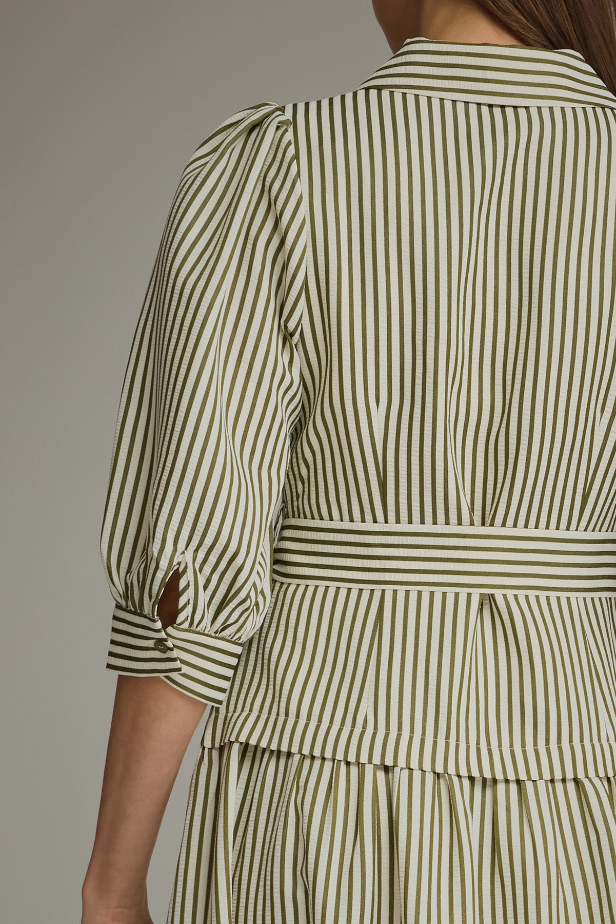 Back view of a woman modeling an olive green and white vertically striped midi dress with three-quarter sleeves and a tie waist and drop waist, against a neutral gray studio background.