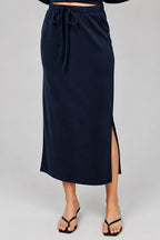 A navy blue midi skirt with a drawstring waist and a side slit, shown on a model wearing black thong sandals against a neutral beige background.