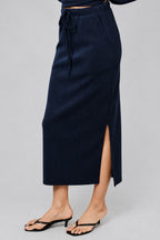 A navy blue midi skirt with a drawstring waist and a side slit, shown on a model wearing black thong sandals against a neutral beige background.