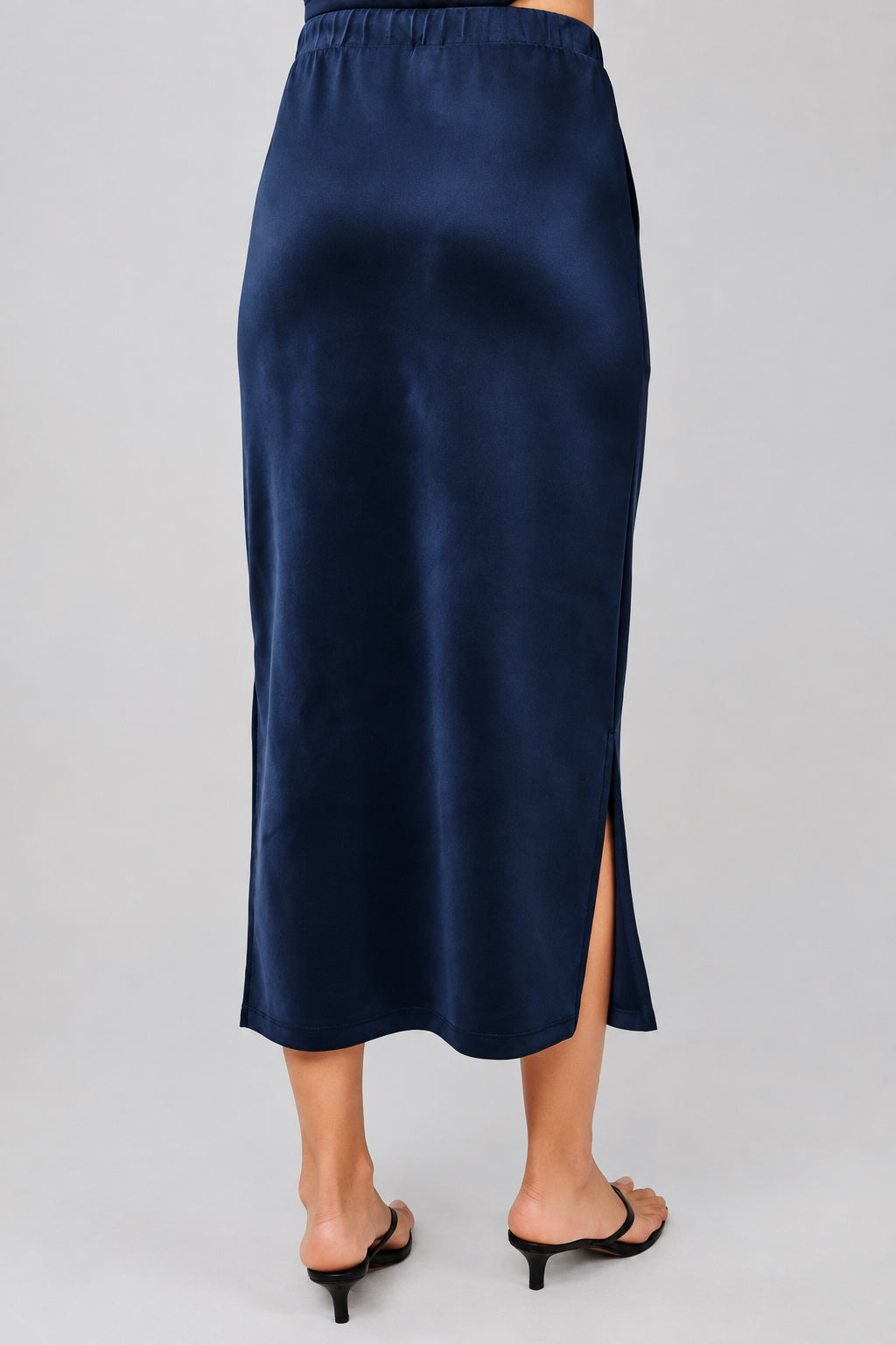 Back view of a navy blue midi skirt with a drawstring waist and a side slit, shown on a model wearing black thong sandals against a neutral beige background.