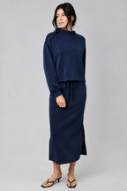 A woman modeling a navy blue two-piece set consisting of a long-sleeve top and a matching midi skirt, styled with black heeled thong sandals against a textured beige studio backdrop.