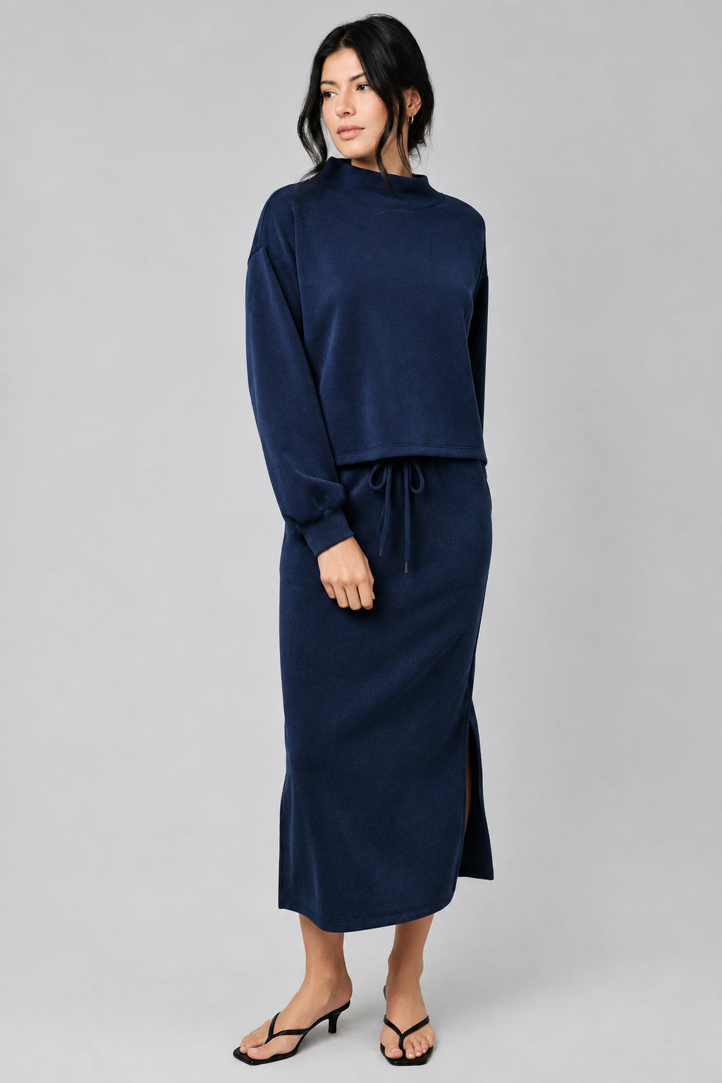 A woman modeling a navy blue two-piece set consisting of a long-sleeve top and a matching midi skirt, styled with black heeled thong sandals against a textured beige studio backdrop.