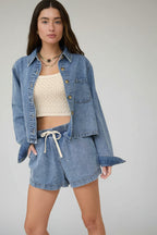 Woman wearing a denim jacket and shorts with a beige top against a neutral background