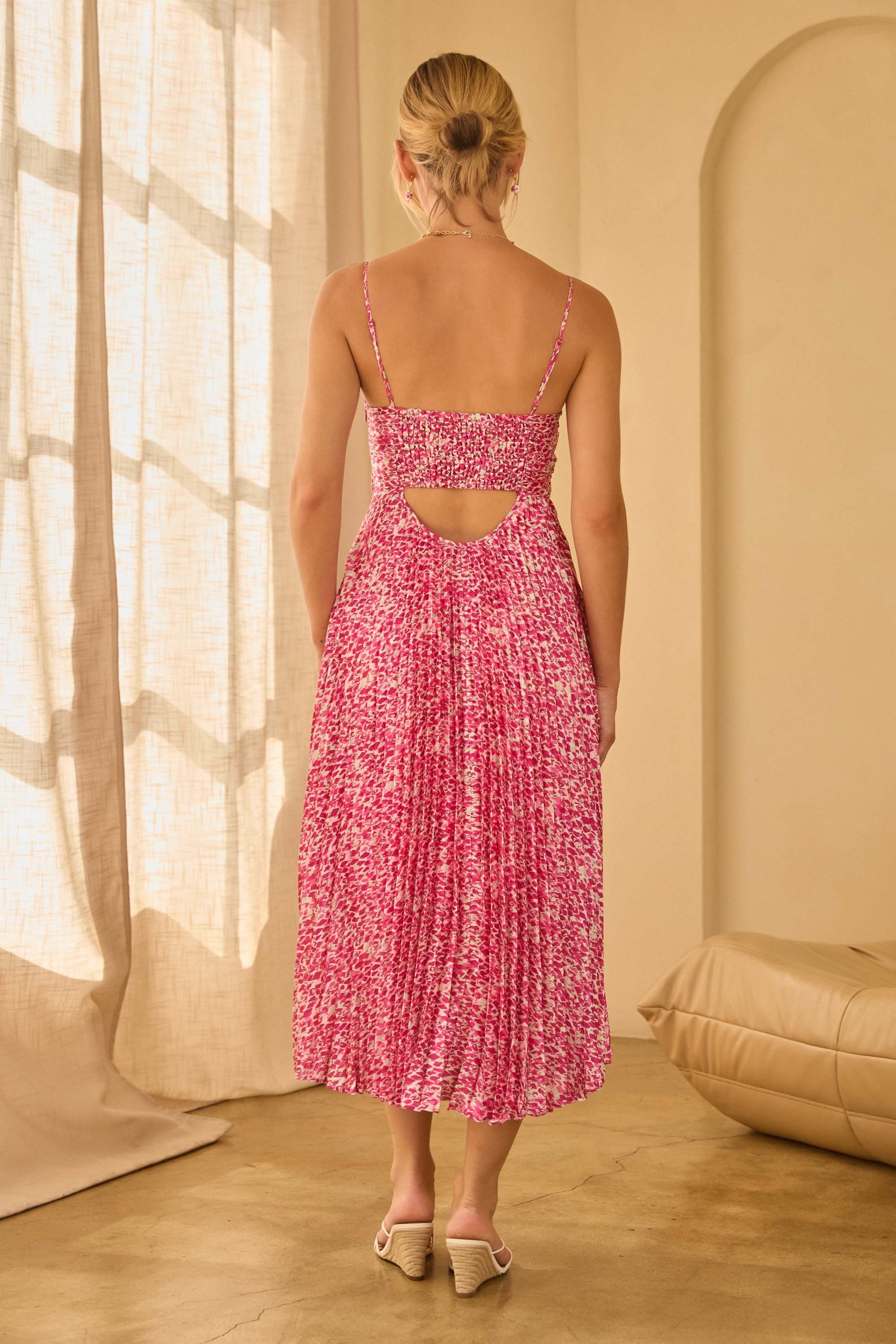Back view of a woman wearing a pink floral spaghetti strap dress with pleated skirt and cut out back in a softly lit room.