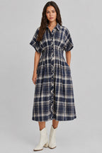 A woman modeling a dolman sleeve button-front midi dress with a navy and cream plaid pattern, paired with white ankle boots, against a plain light beige studio background.