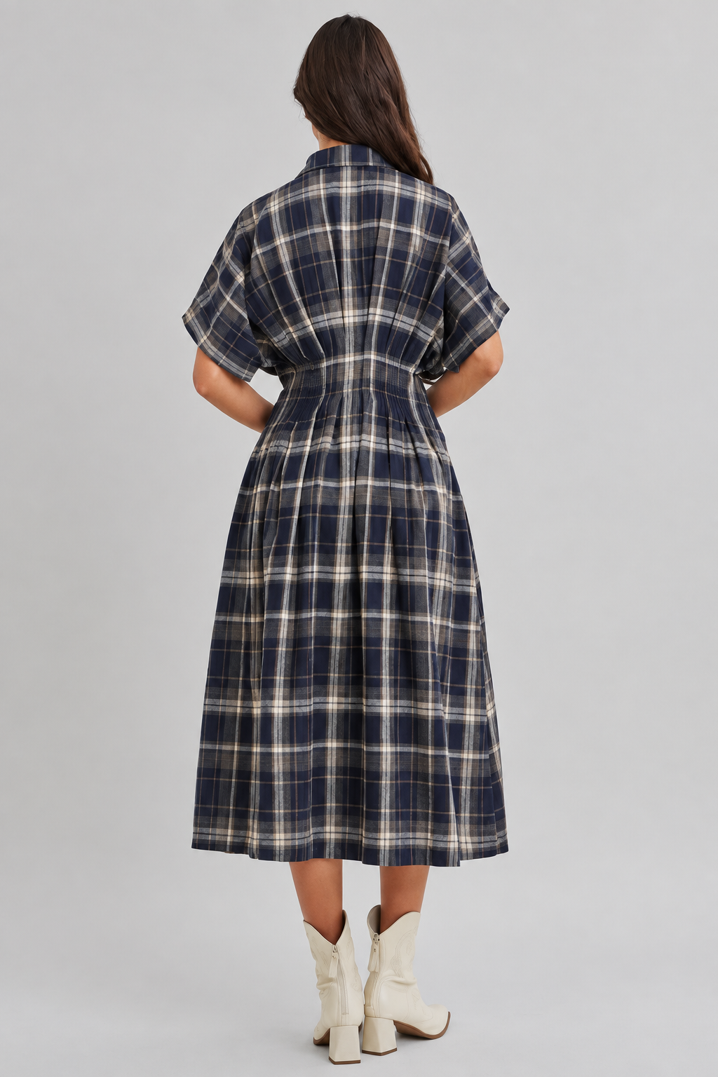 Back view of a woman modeling a dolman sleeve button-front midi dress with a navy and cream plaid pattern, paired with white ankle boots, against a plain light beige studio background.