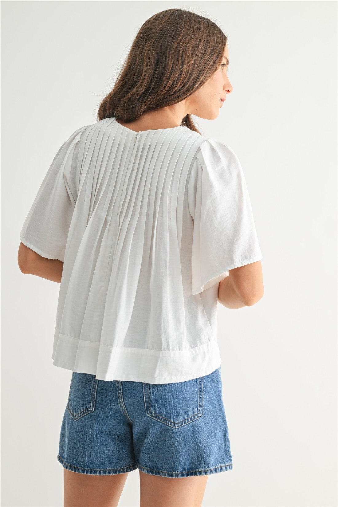 Back view of a woman wearing a white pin tuck top and blue jeans against a plain background