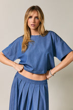 A woman wearing a cropped short-sleeve top and matching pleated skirt against a plain light gray studio background.